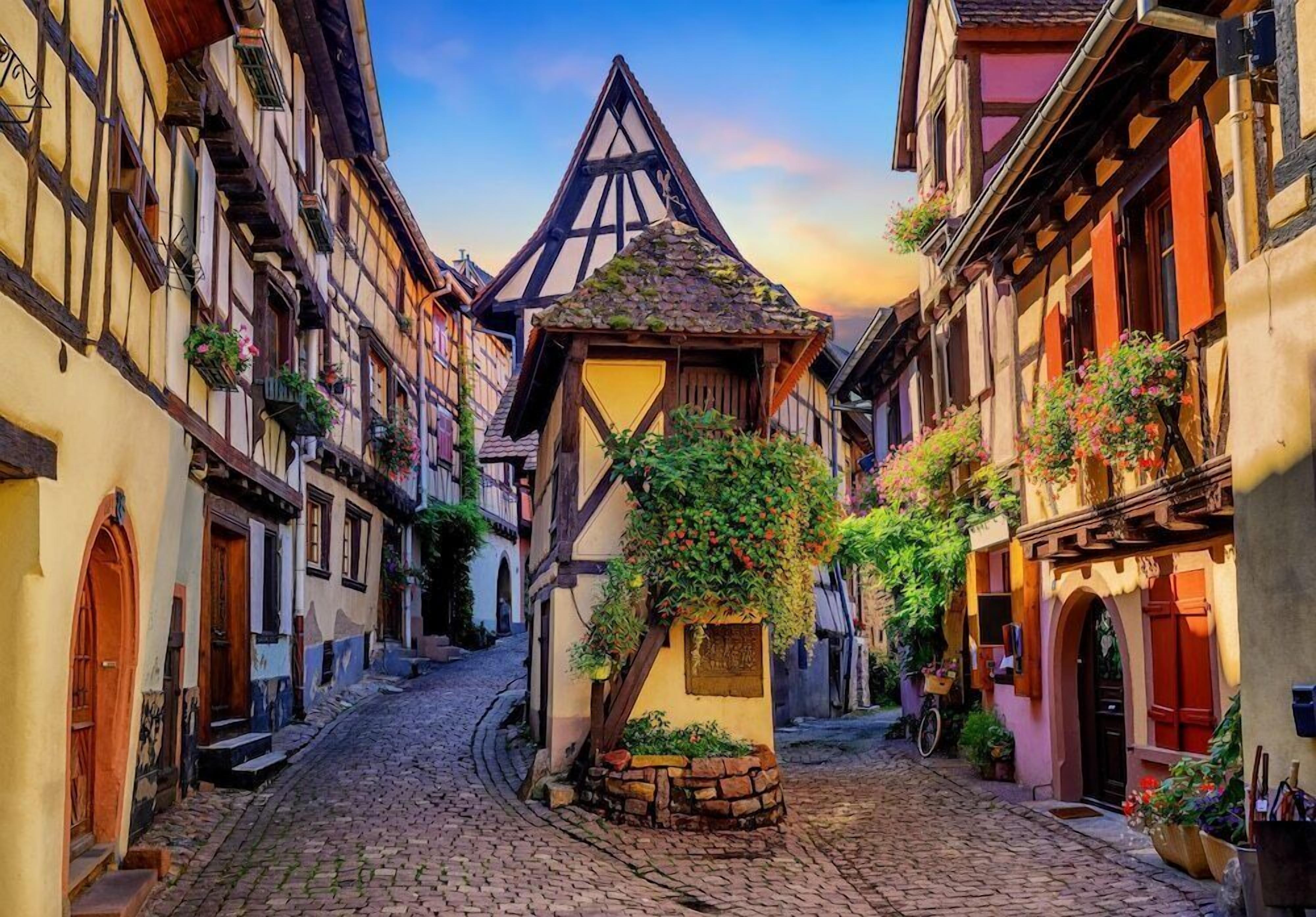 Une ruelle étroite pavée, bordée de bâtiments traditionnels à colombages, avec une structure centrale surmontée d’un toit triangulaire et des jardinières aux fenêtres.