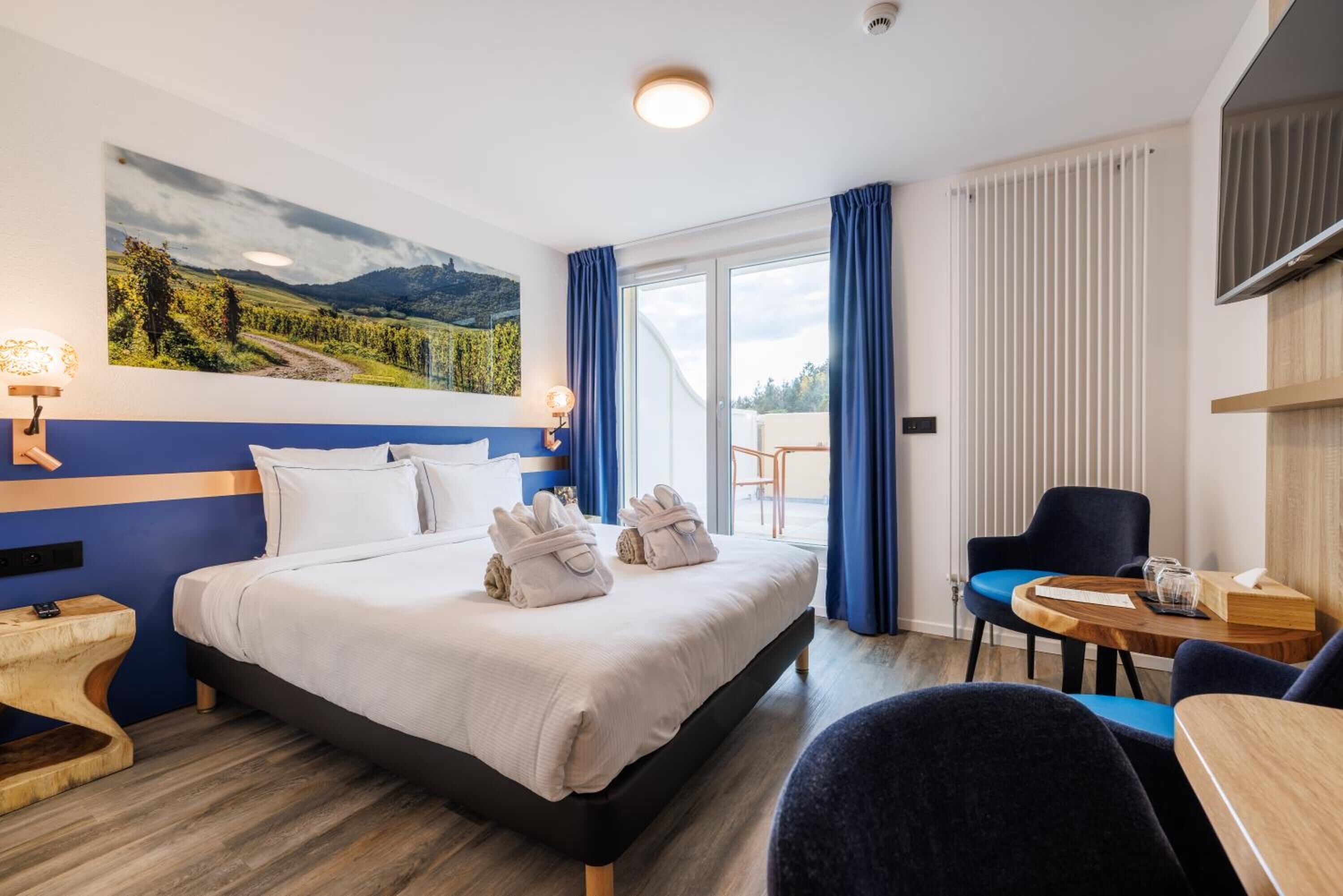 Une chambre d’hôtel avec un lit, un bureau, une chaise, une fenêtre avec des rideaux et un tableau au mur.