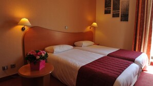 Une chambre d’hôtel avec un lit double, deux lampes de chevet, une petite table sur laquelle se trouve une boîte cadeau rose et des fleurs, ainsi que deux œuvres d’art murales.