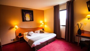 Une chambre d’hôtel avec un grand lit, deux lampes de chevet, un tableau au mur, un bureau avec une lampe et une fenêtre avec des rideaux.