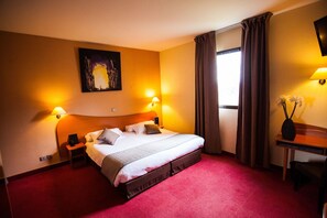 Habitación de hotel con una cama grande, dos lámparas de noche, un cuadro en la pared, un escritorio con lámpara y una ventana con cortinas.