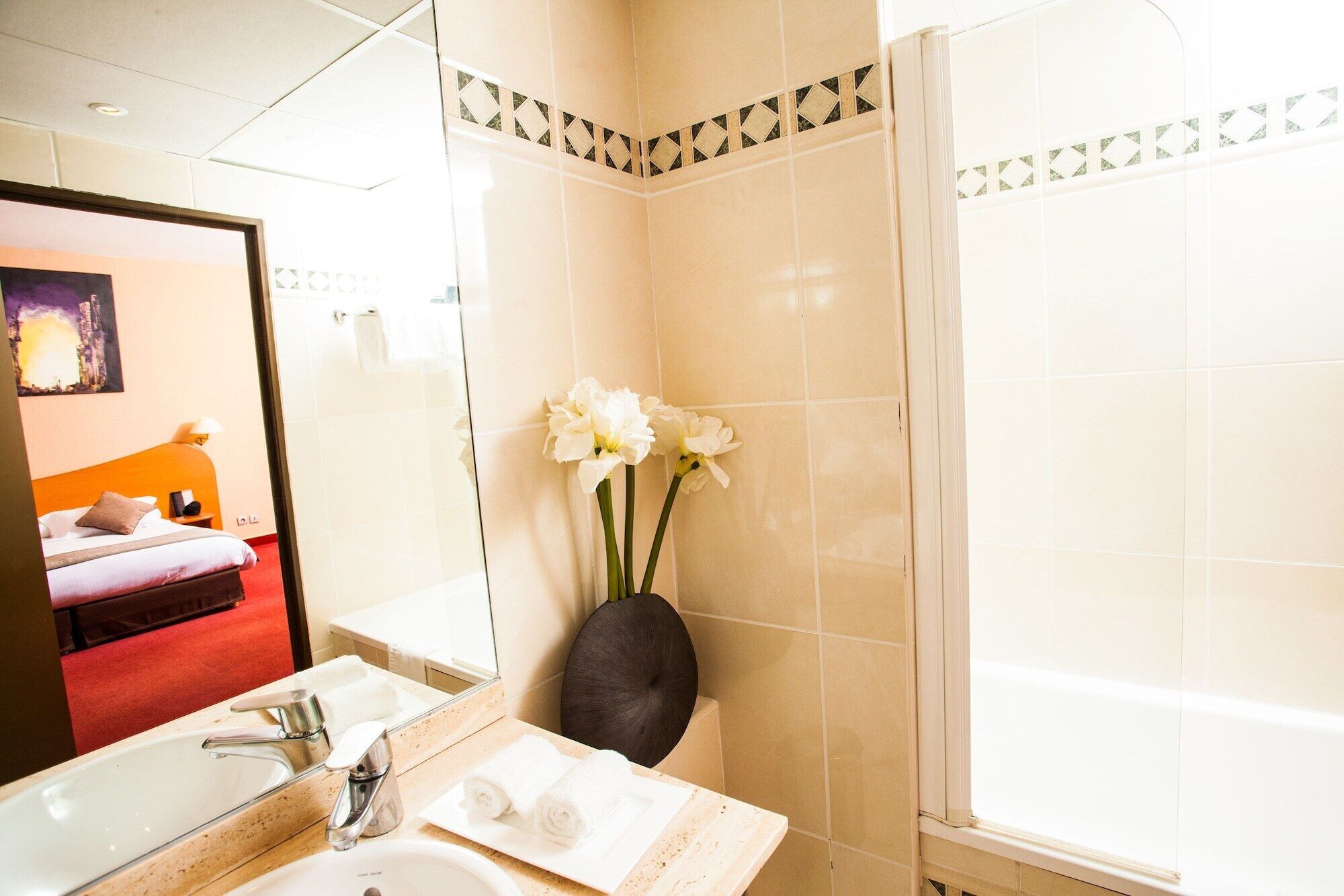 Une salle de bain d’hôtel avec un grand miroir, un lavabo, une baignoire et un vase rempli de fleurs.