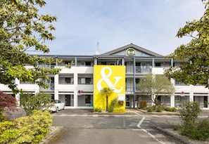 A hotel with a large yellow sign featuring a white anchor symbol, multiple floors, and a parking area with a white car. - B&B Hotel Rennes Nord St Gregoire (Saint-Gregoire)