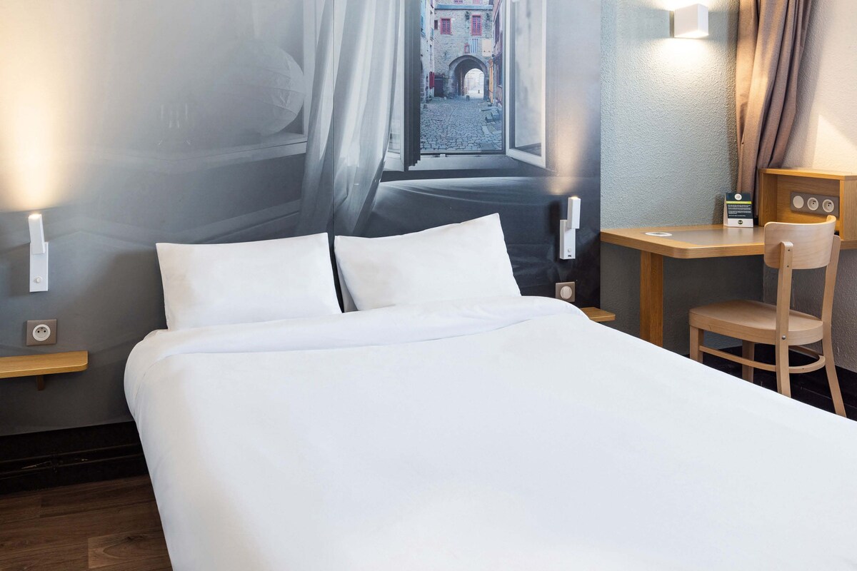 A neatly made bed with white linens in a hotel room. There is a wooden desk with a chair and a small table lamp. A wall-mounted light fixture is visible on the left side of the bed.