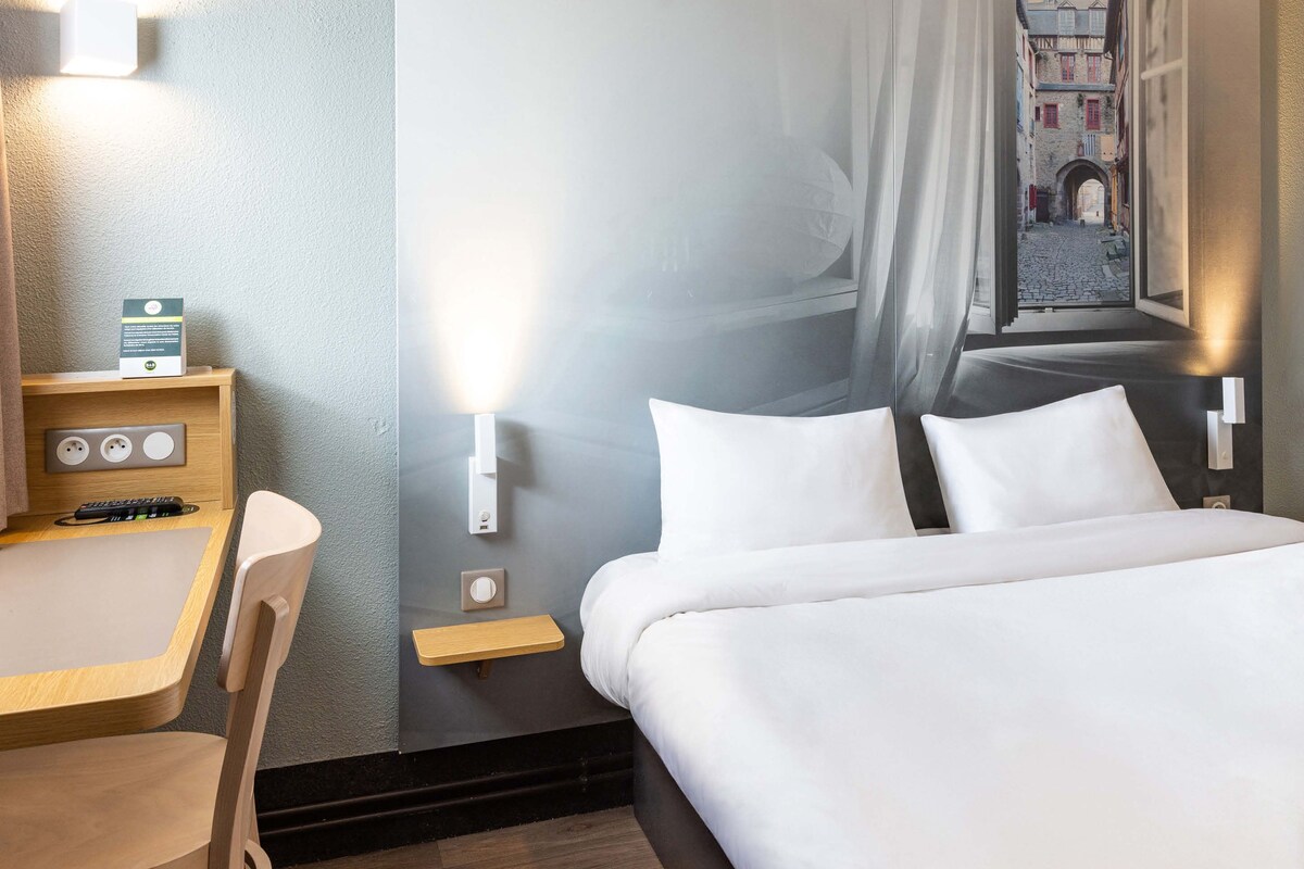A hotel room with a bed, a desk, a chair, and a view of a cobblestone street through a window.