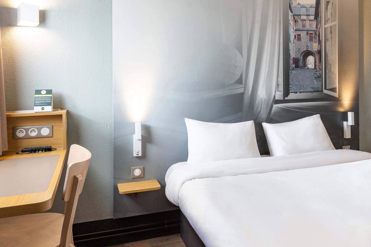 A hotel room with a bed, a desk, a chair, and a view of a cobblestone street through a window.