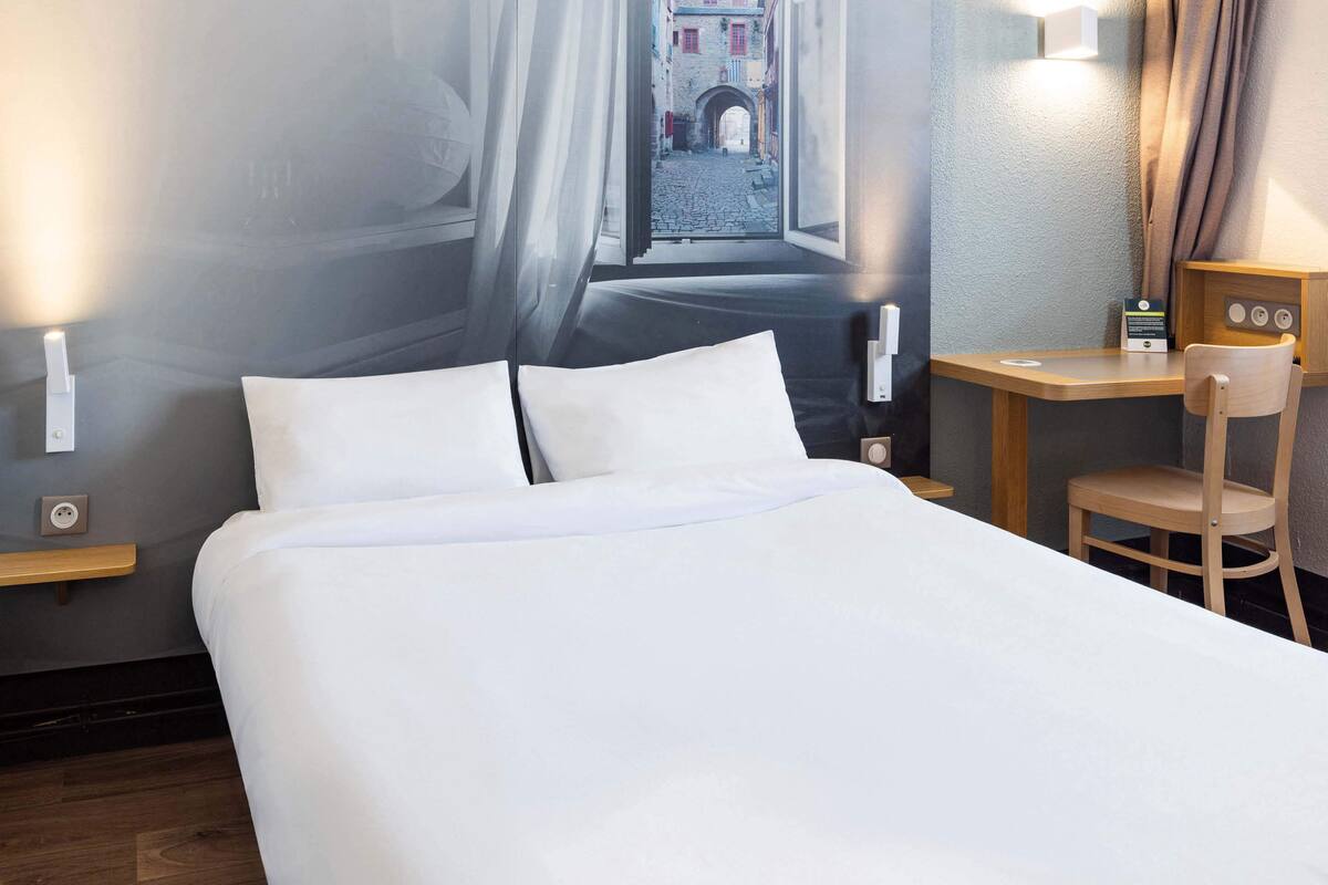 A neatly made bed with white linens in a hotel room. There is a wooden desk with a chair and a small table lamp. A wall-mounted light fixture is visible on the left side of the bed.