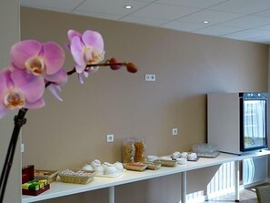Un espace d’accueil d’hôtel avec un comptoir où sont exposés des produits pour le petit-déjeuner, un téléviseur et un vase contenant des orchidées.