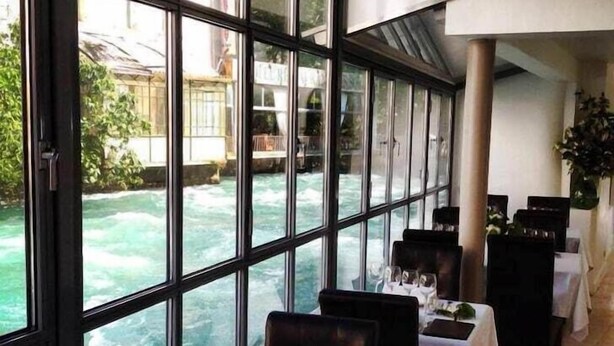 Un restaurant avec des tables dressées pour le service, de grandes fenêtres et une vue sur une rivière.