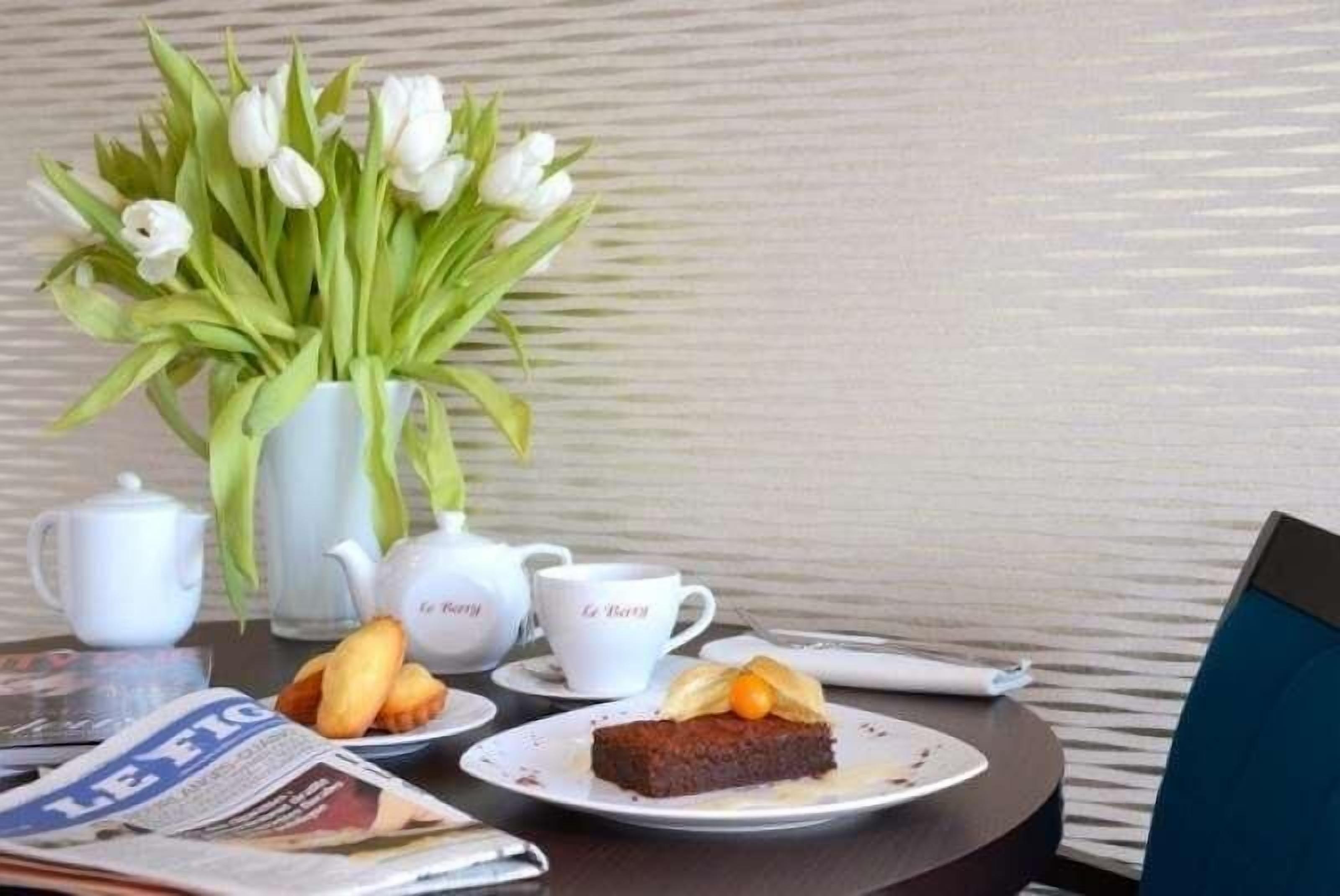 A table with a vase of white tulips, a teapot, a cup, a plate with a dessert, and a newspaper.