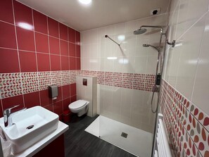 Een moderne badkamer met een rood-wit betegeld ontwerp, een wit wastafelmeubel, een toilet en een douchegedeelte.