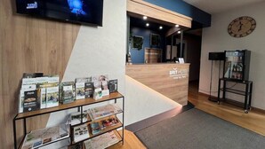 Een hotel receptie met een houten balie, een wandtelevisie, een klok en een presentatie van brochures en kaarten.