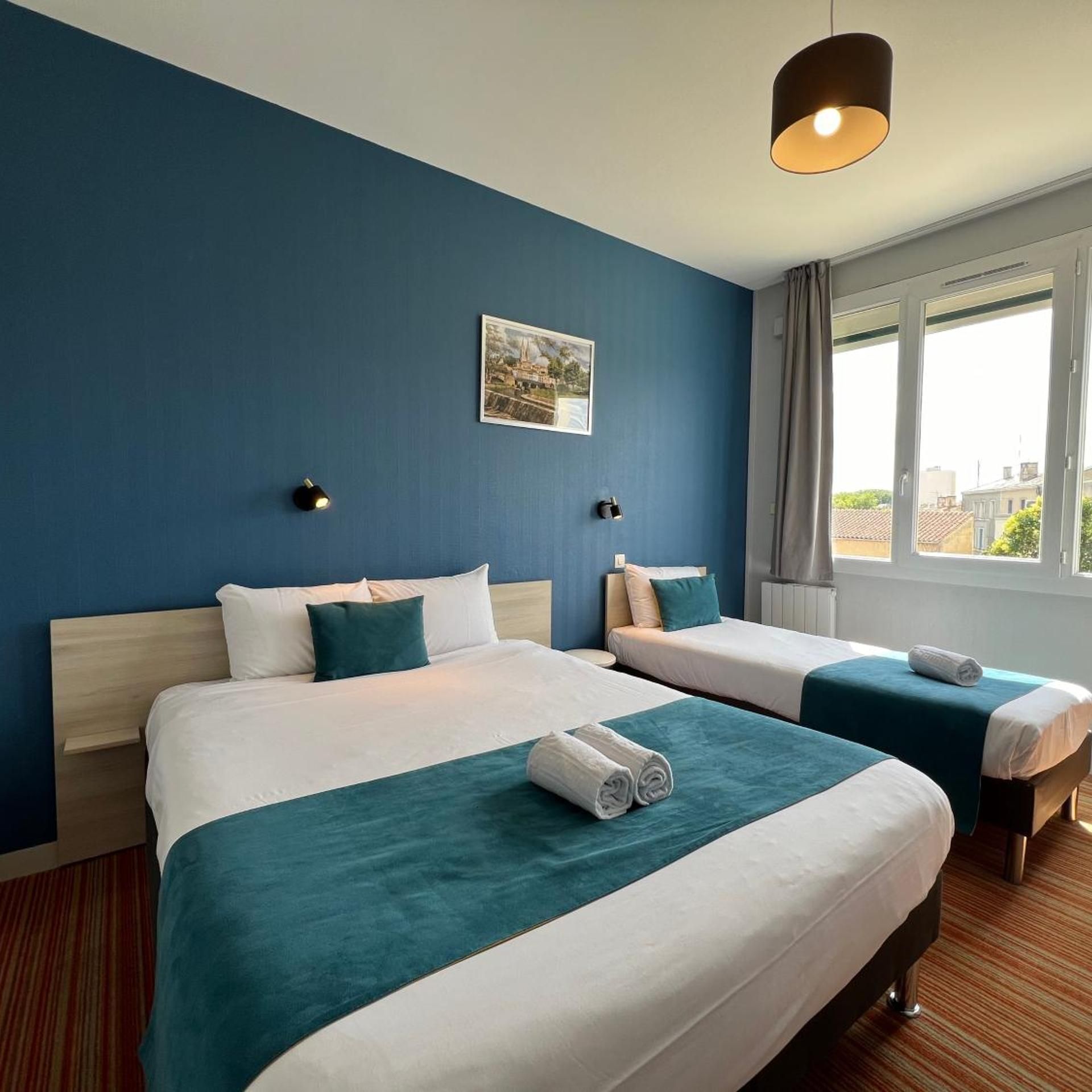 A hotel room with two beds, a large window, a painting on the wall, and a ceiling light.