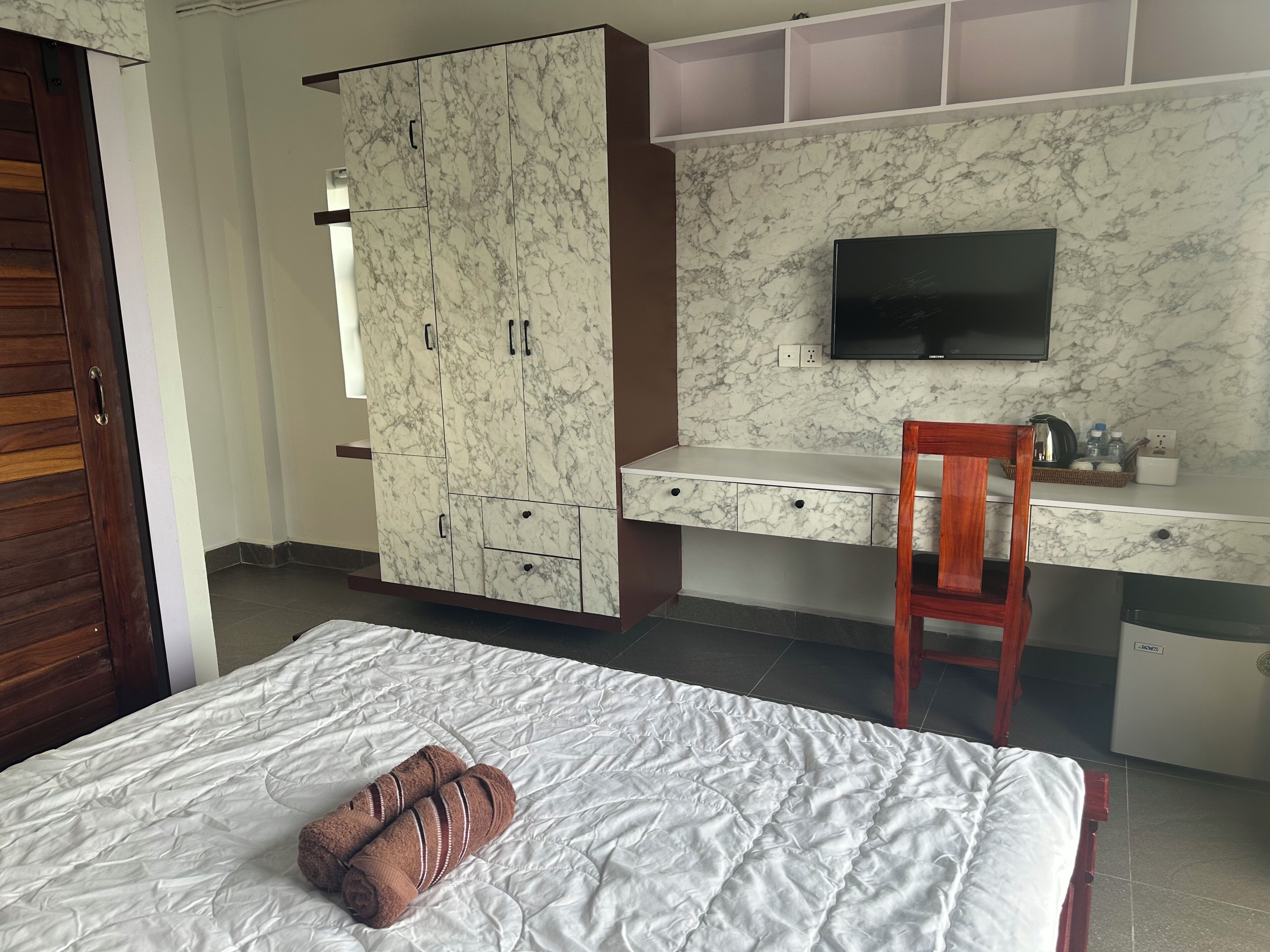 A bedroom with a bed, a wardrobe, a desk with a chair, a television, and a small kitchen area.