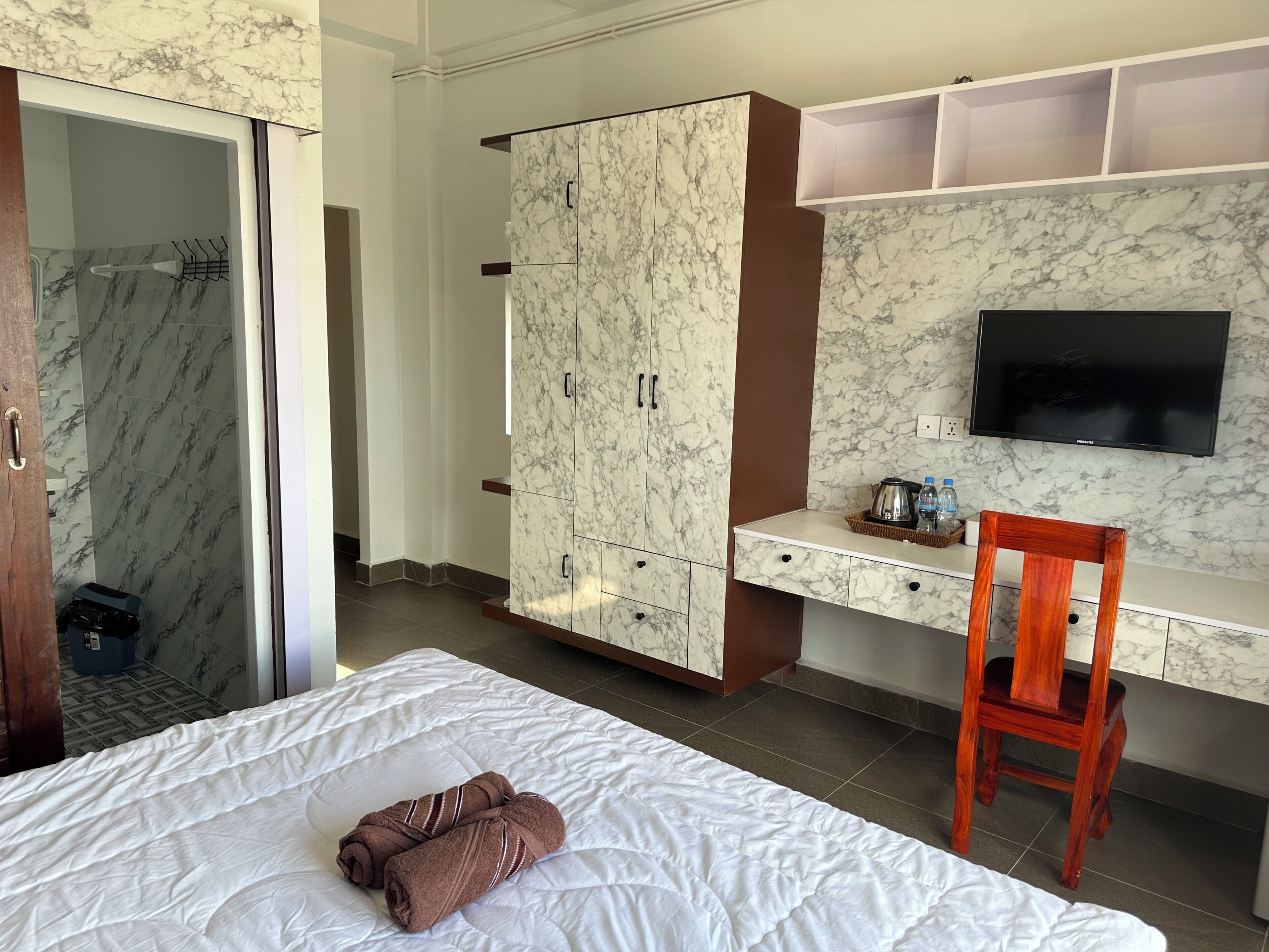 A hotel room with a bed, a wooden chair, a desk with a TV, a wardrobe, and a shower area.
