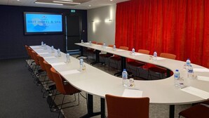 Konferenssihuone, jossa on pitkä pöytä, oranssit tuolit ja punainen verho taustalla. Näytöllä näkyy teksti: ”BRIT HOTEL & SPA”.