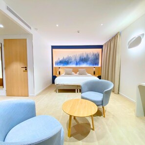 Una habitación de hotel moderna con una cama, dos sillones, una mesita de madera y una obra de arte colgada en la pared.