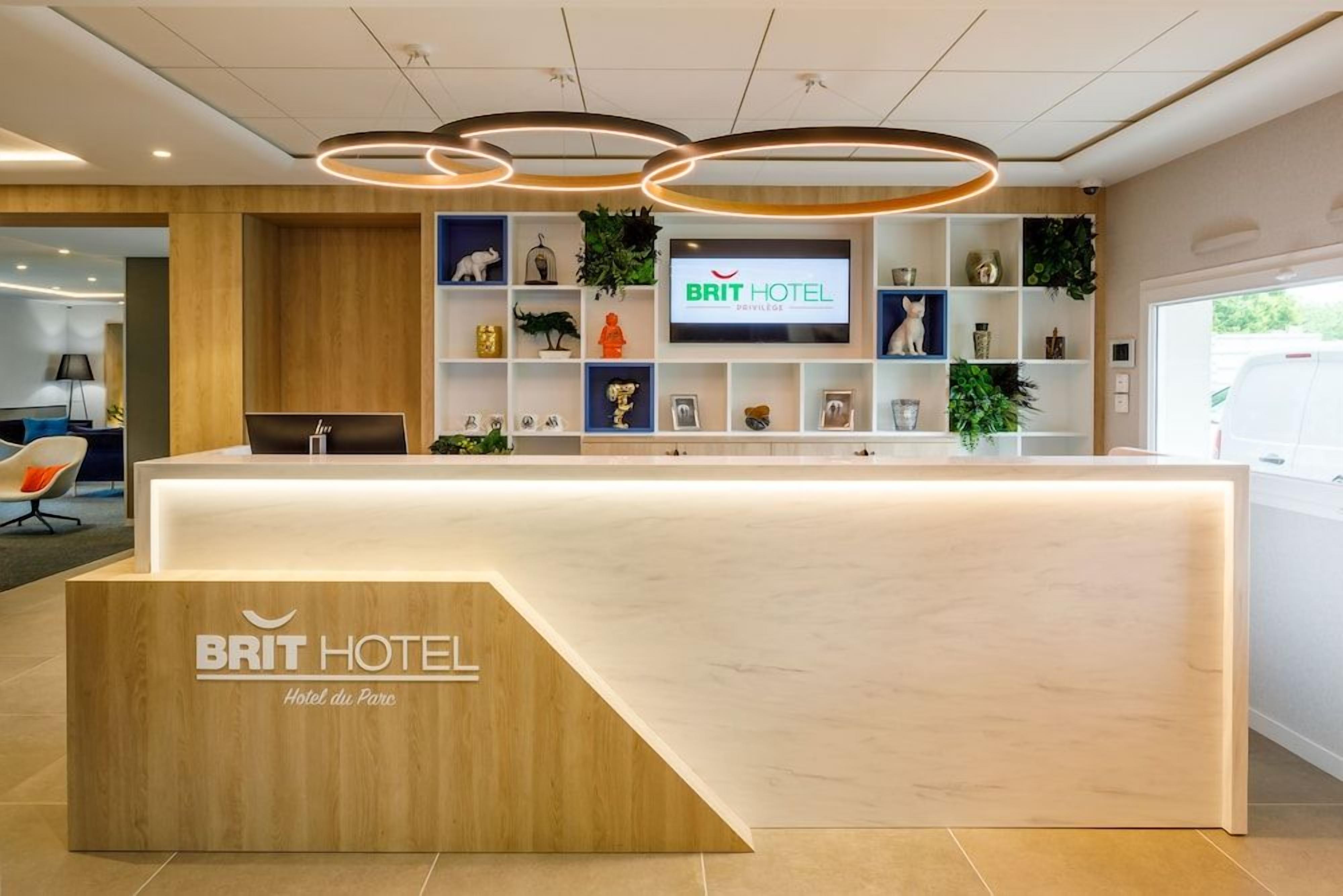 Un espace d’accueil doté d’un comptoir de réception en bois, d’un écran de télévision affichant le nom de l’hôtel, et d’étagères contenant divers objets et plantes.