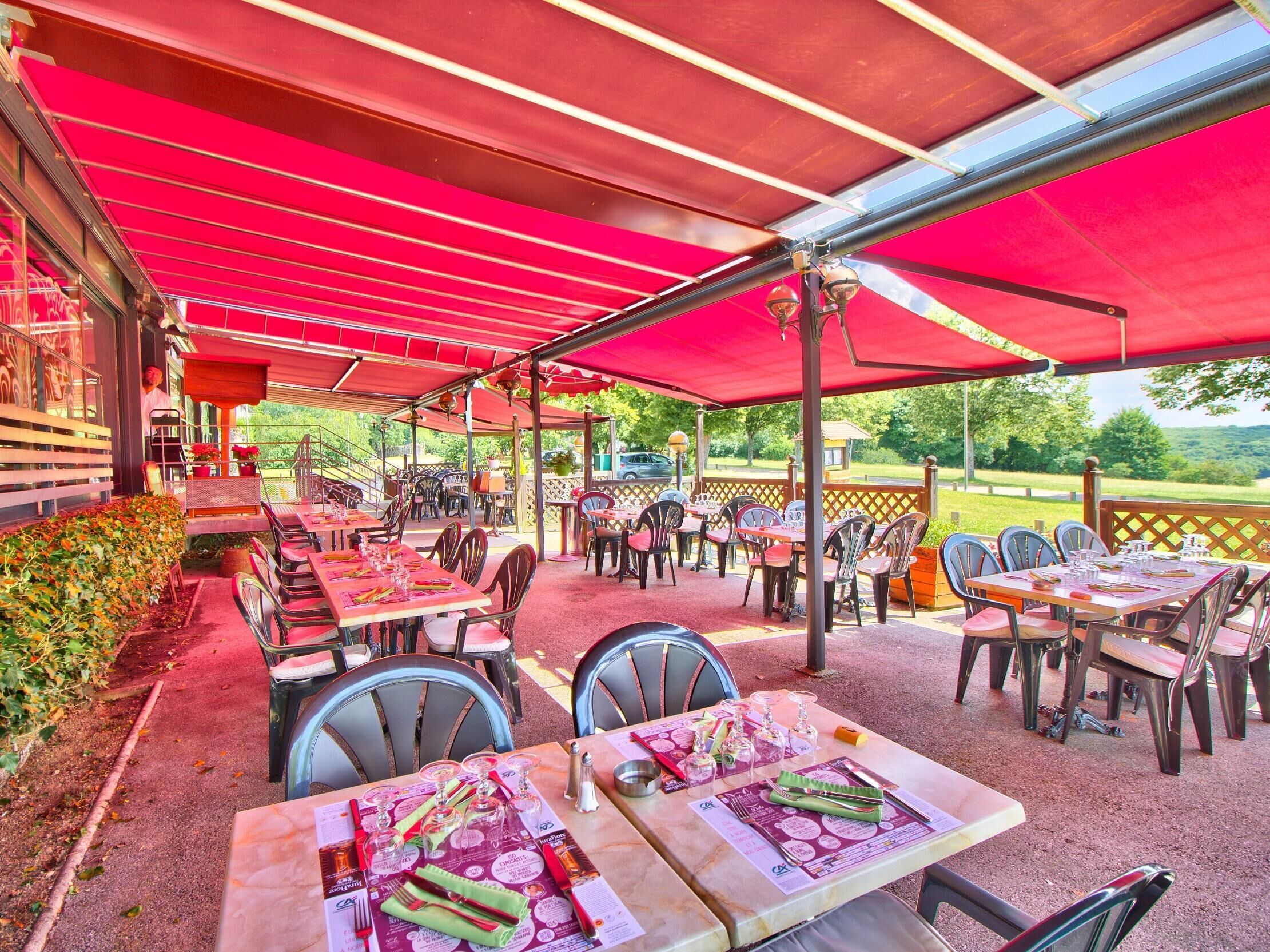 Een terras met tafels en stoelen onder rode parasols, omringd door groen en uitzicht op het platteland.