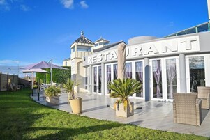 Un restaurante con fachada moderna, amplias ventanas y terraza amueblada con mobiliario de mimbre.