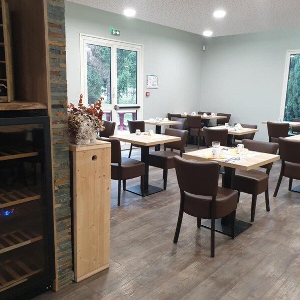 A dining area with multiple tables and chairs, a wooden counter, and a view of the outdoors through large windows. - Hotel Des Portes De Meuse (Pagny-sur-Meuse)