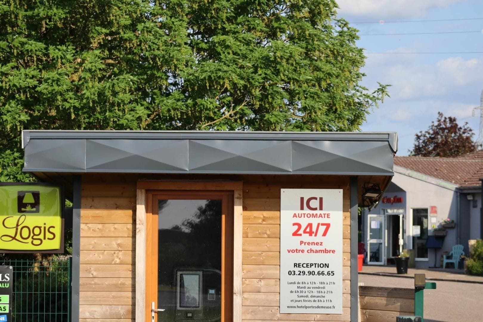 Un petit bâtiment moderne avec une façade en bois et un toit gris. Il porte une enseigne indiquant un service de réception automatisé disponible 24 heures sur 24, 7 jours sur 7, ainsi qu'un numéro de téléphone à contacter.