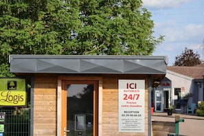 A small, modern building with a wooden facade and a grey roof. It has a sign indicating 24/7 automated reception and a contact number. - Hotel Des Portes De Meuse (Pagny-sur-Meuse)