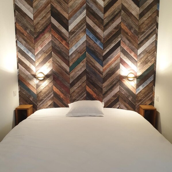 A bed with a white mattress and pillows, flanked by two nightstands with lamps, against a wall with a chevron-patterned wooden panel. - Hotel Des Portes De Meuse (Pagny-sur-Meuse)