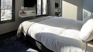 A modern hotel room with a large bed, a TV, a mirror, and a window with a city view.