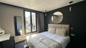 A modern hotel room with a large bed, a desk, a round mirror, and a view of the city through a large window.
