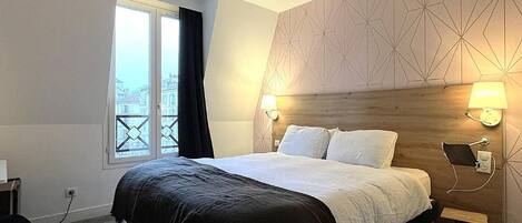 A hotel room with a bed, bedside lamps, a window with curtains, and a geometric-patterned wall.
