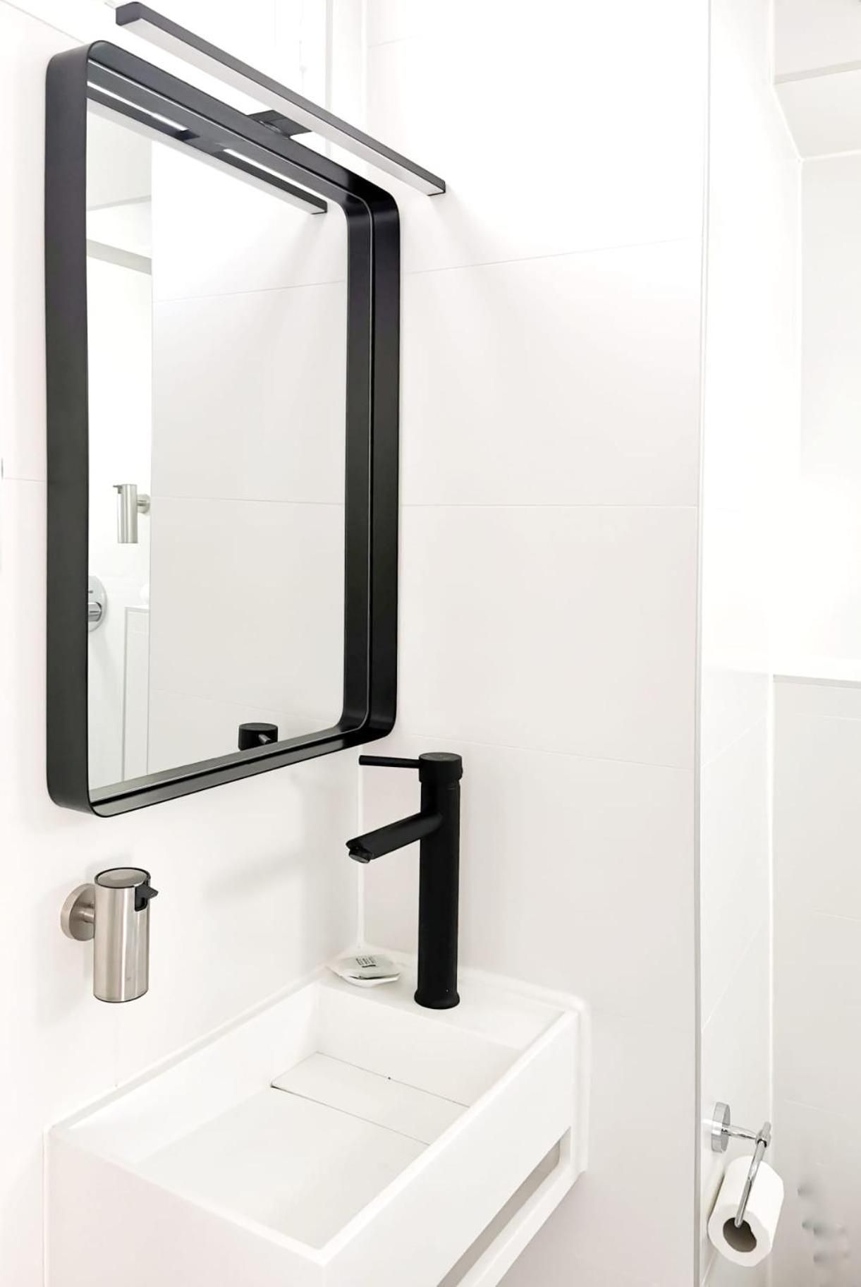 Une salle de bain moderne avec un lavabo blanc, un robinet noir, un porte-gobelet en acier inoxydable et un miroir rectangulaire à cadre noir.