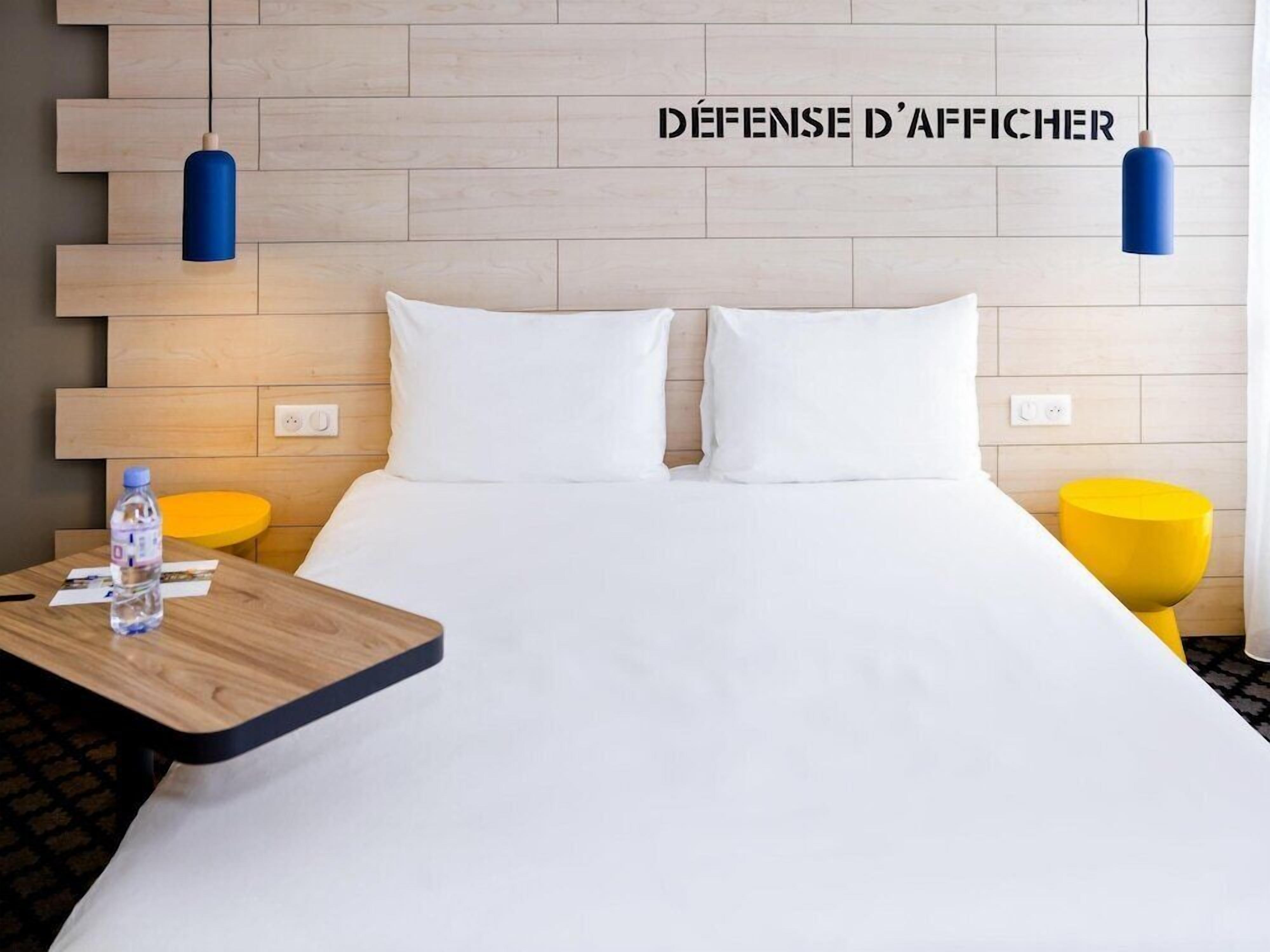 A hotel room with a bed, a wooden nightstand, a table with a water bottle, and two pendant lights.