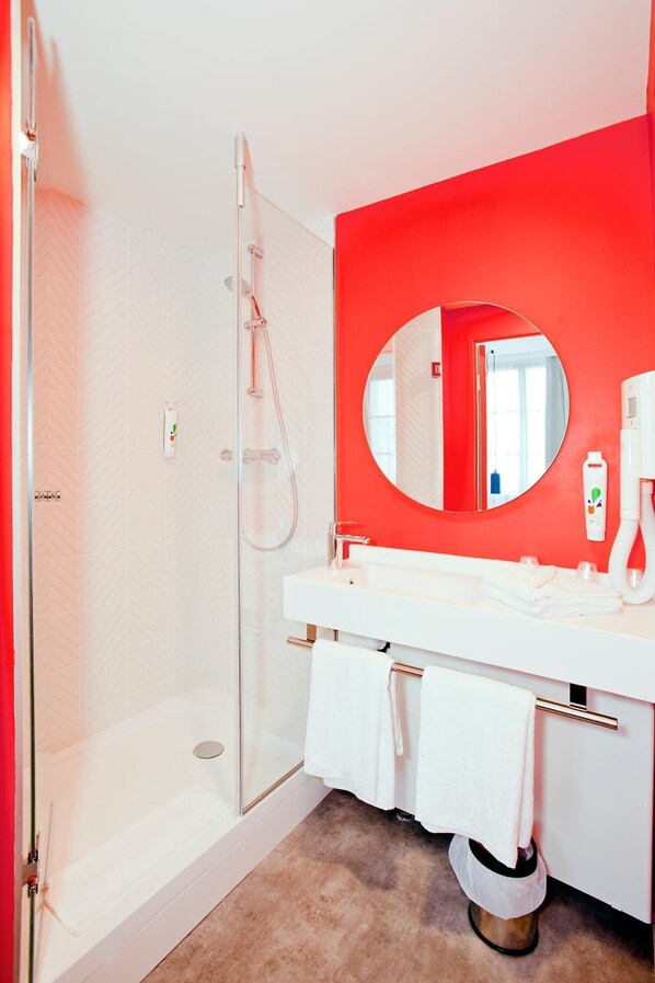 A modern bathroom with a glass-enclosed shower, a white vanity with a round mirror, and a red accent wall. - Ibis Styles Chlons-en-champagne Centre (Chalons-en-Champagne)
