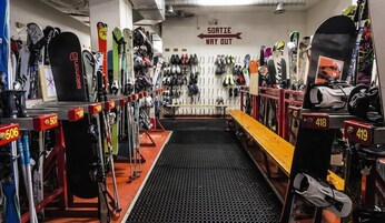 A ski equipment rental shop with rows of skis and snowboards, a bench, and a sign indicating the way out.