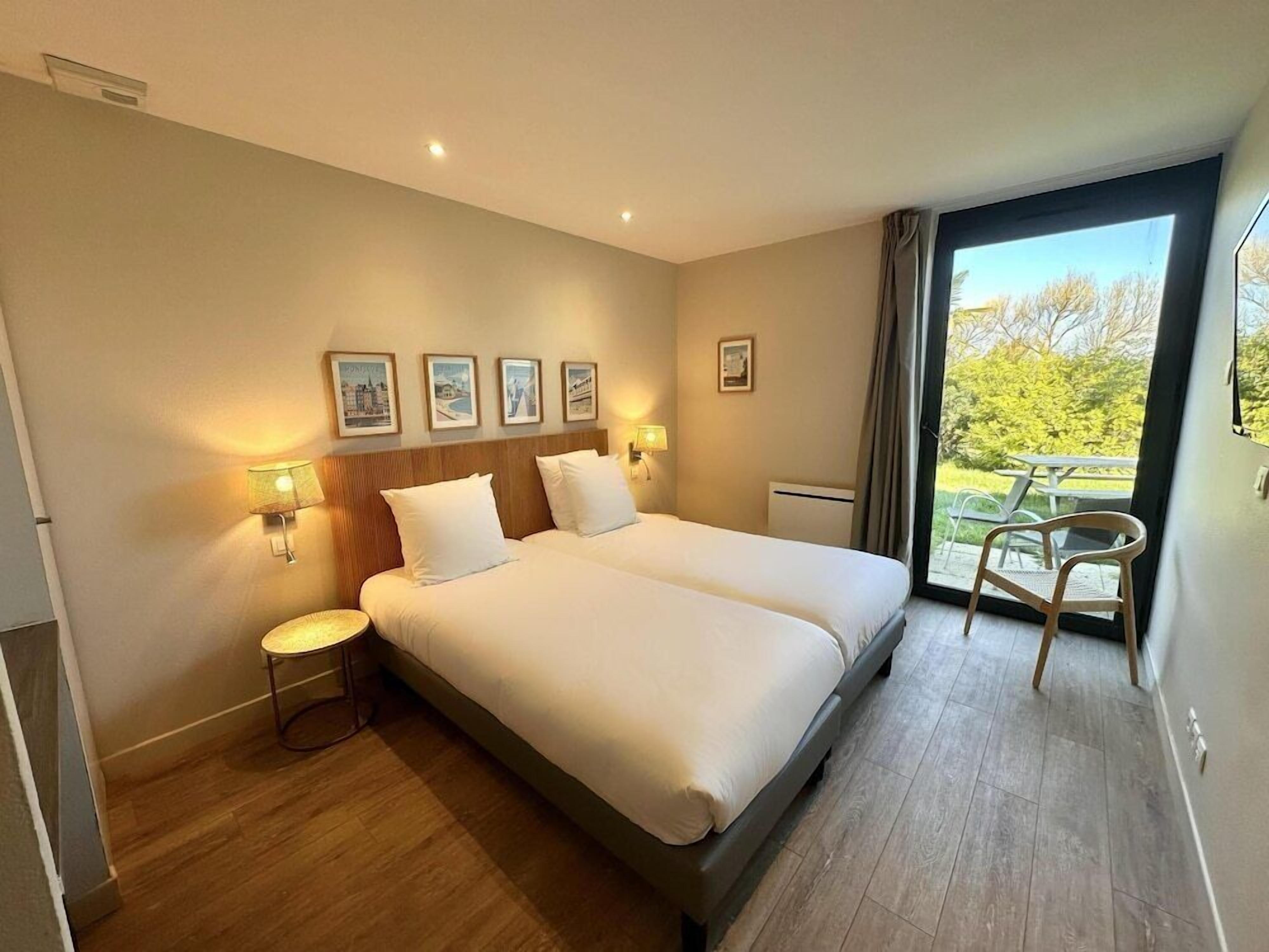 A hotel room with a large bed, two bedside tables with lamps, a window with a view of greenery, and a small table with chairs outside.