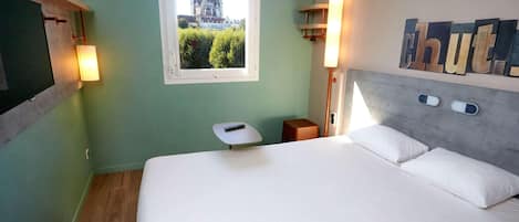 A hotel room with a bed, a window with a view of a building, a bedside table, and a wall-mounted light fixture.