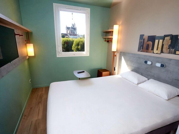 Un cuarto de hotel con una cama, una ventana que da a una vista de un edificio, una mesita de noche y una lámpara fijada a la pared.