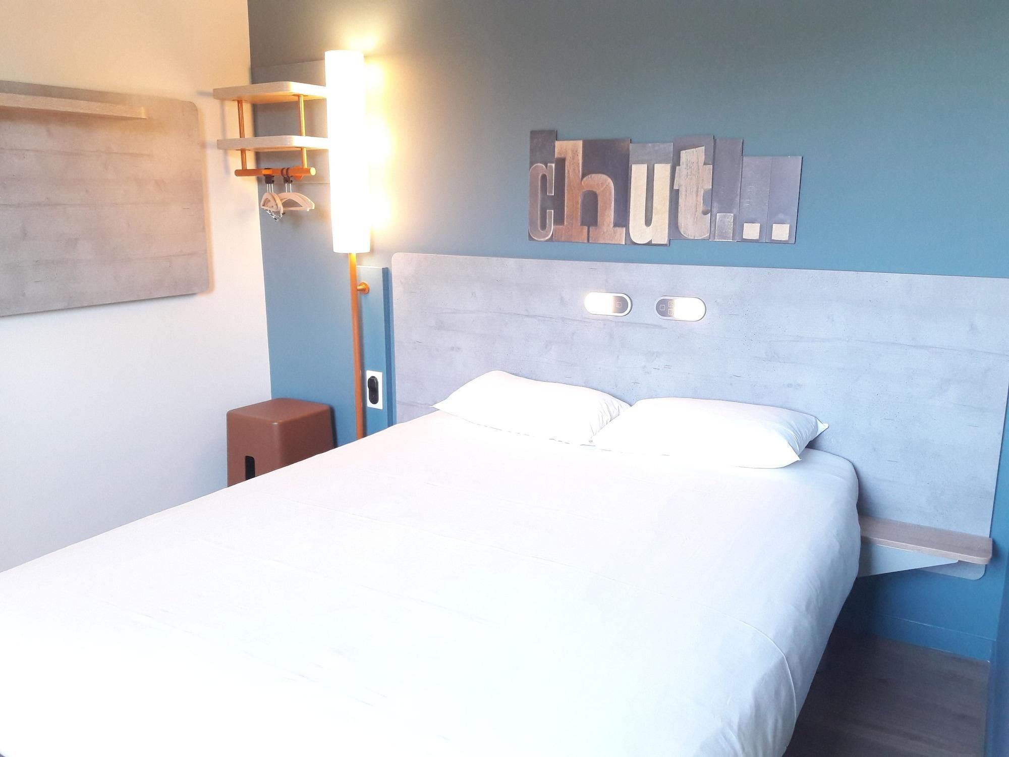A hotel room with a bed, a nightstand, a wall-mounted light, and a sign with the word 'chut.'