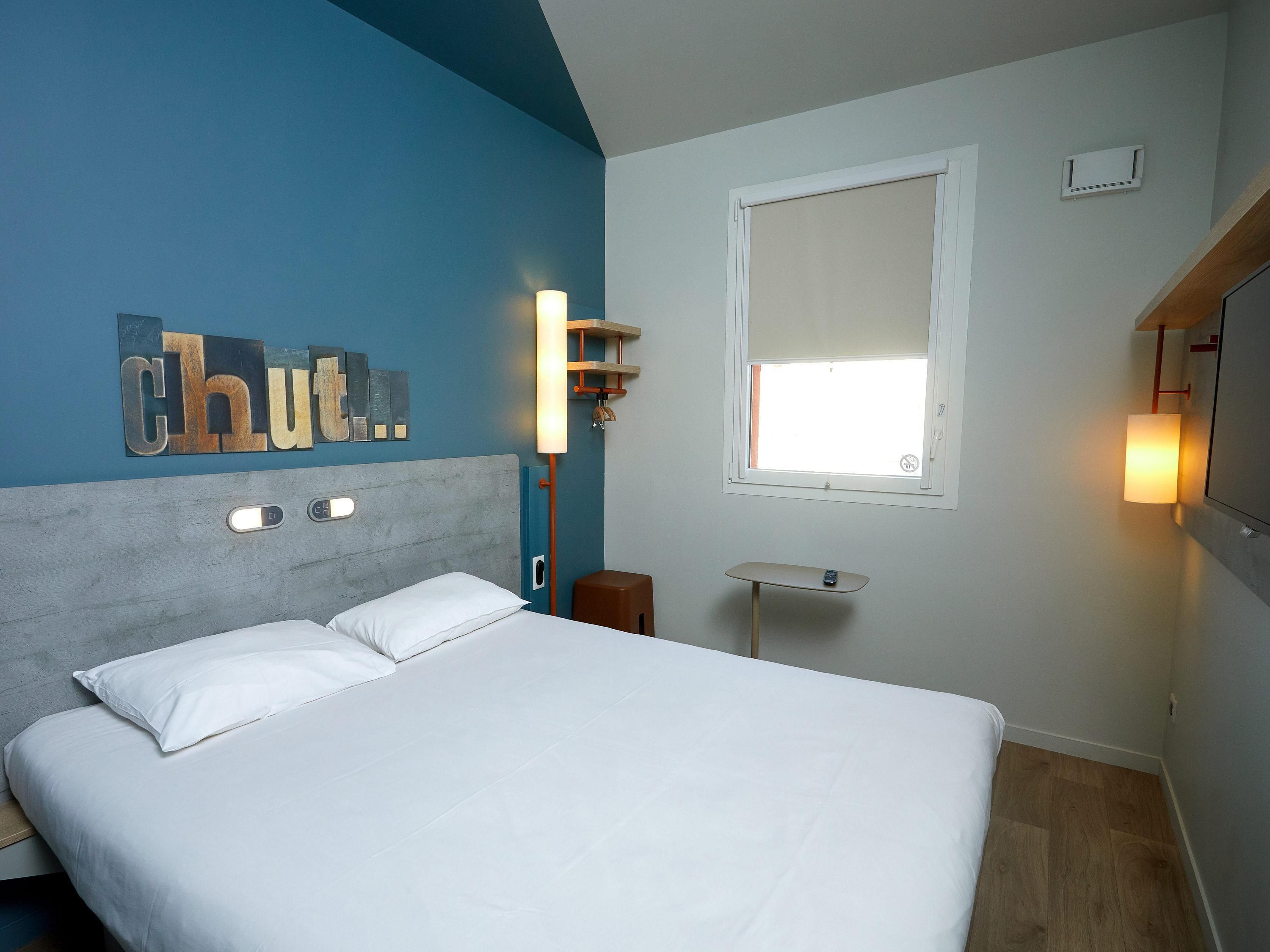 Une chambre d’hôtel avec un lit, une lampe fixée au mur, une fenêtre et un élément décoratif mural.
