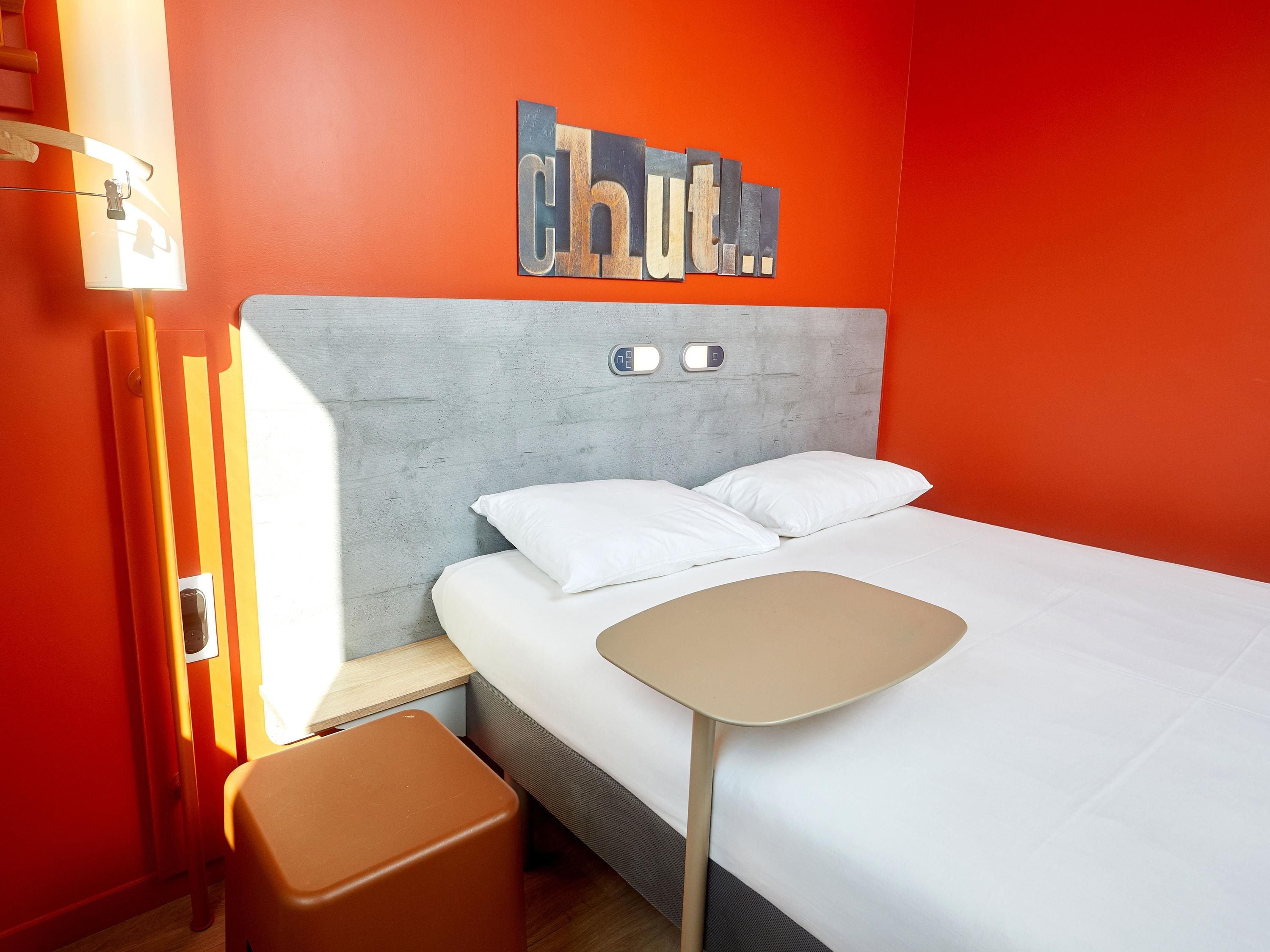 Quarto de hotel moderno com uma cama, uma mesinha, um banquinho e um candeeiro fixo na parede.