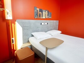 A modern hotel room with a bed, a small table, a stool, and a wall-mounted light fixture.