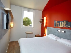 A hotel room with a bed, a TV mounted on the wall, a window with a view of trees, and a red accent wall. - Ibis Budget Auxerre Centre (Auxerre)