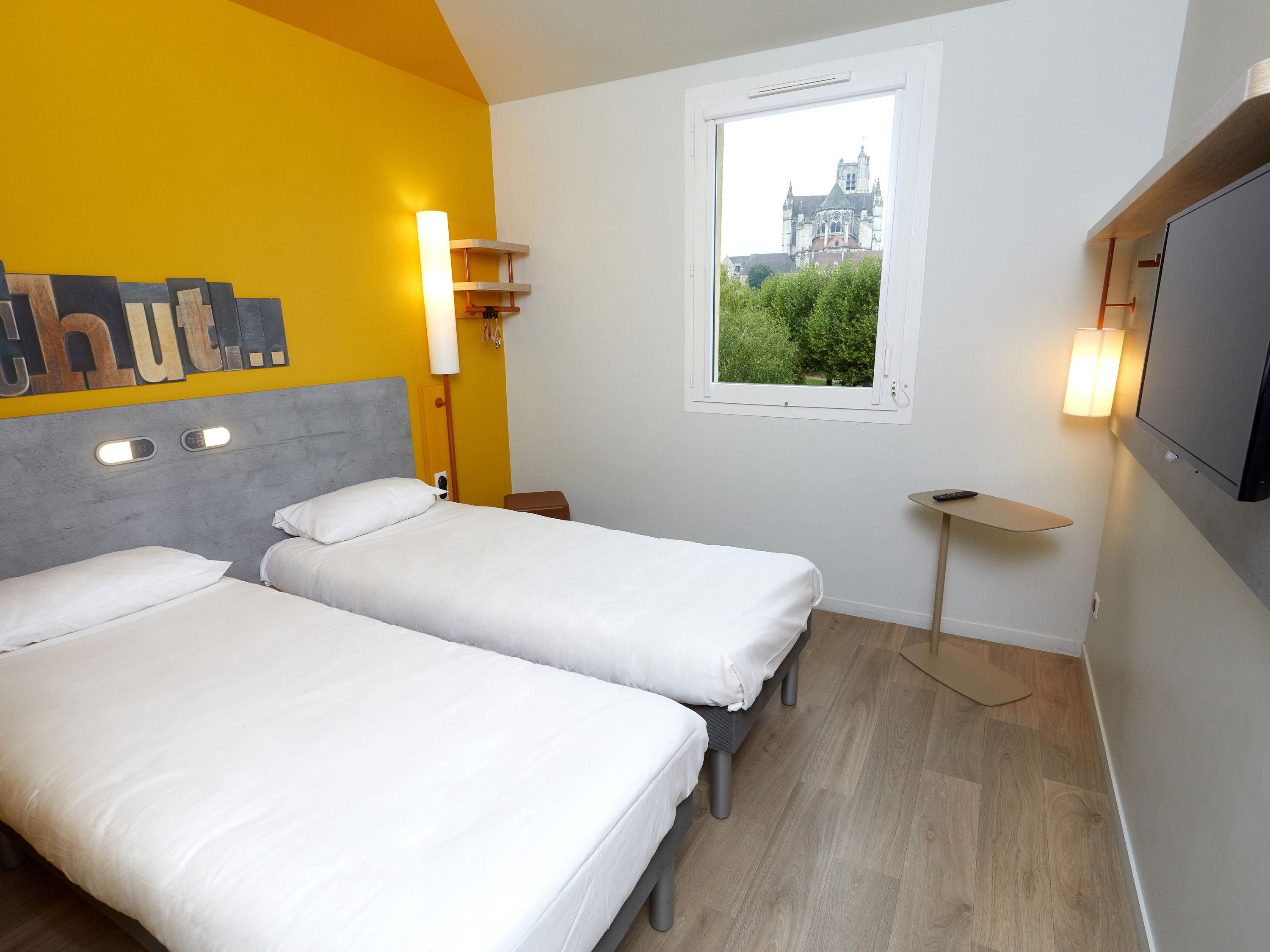 A hotel room with two beds, a window with a view of a building, a TV, and a small table.