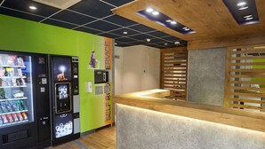 A self-service kiosk area with a vending machine, wooden counter, and a wall with a service sign. - Ibis Budget Auxerre Centre (Auxerre)