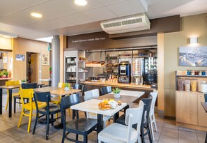 A modern café with tables and chairs, a kitchen area, and a view of a bridge. - B&B Hôtel Lyon Saint-bonnet Mi-plaine (Saint-Bonnet-de-Mure)