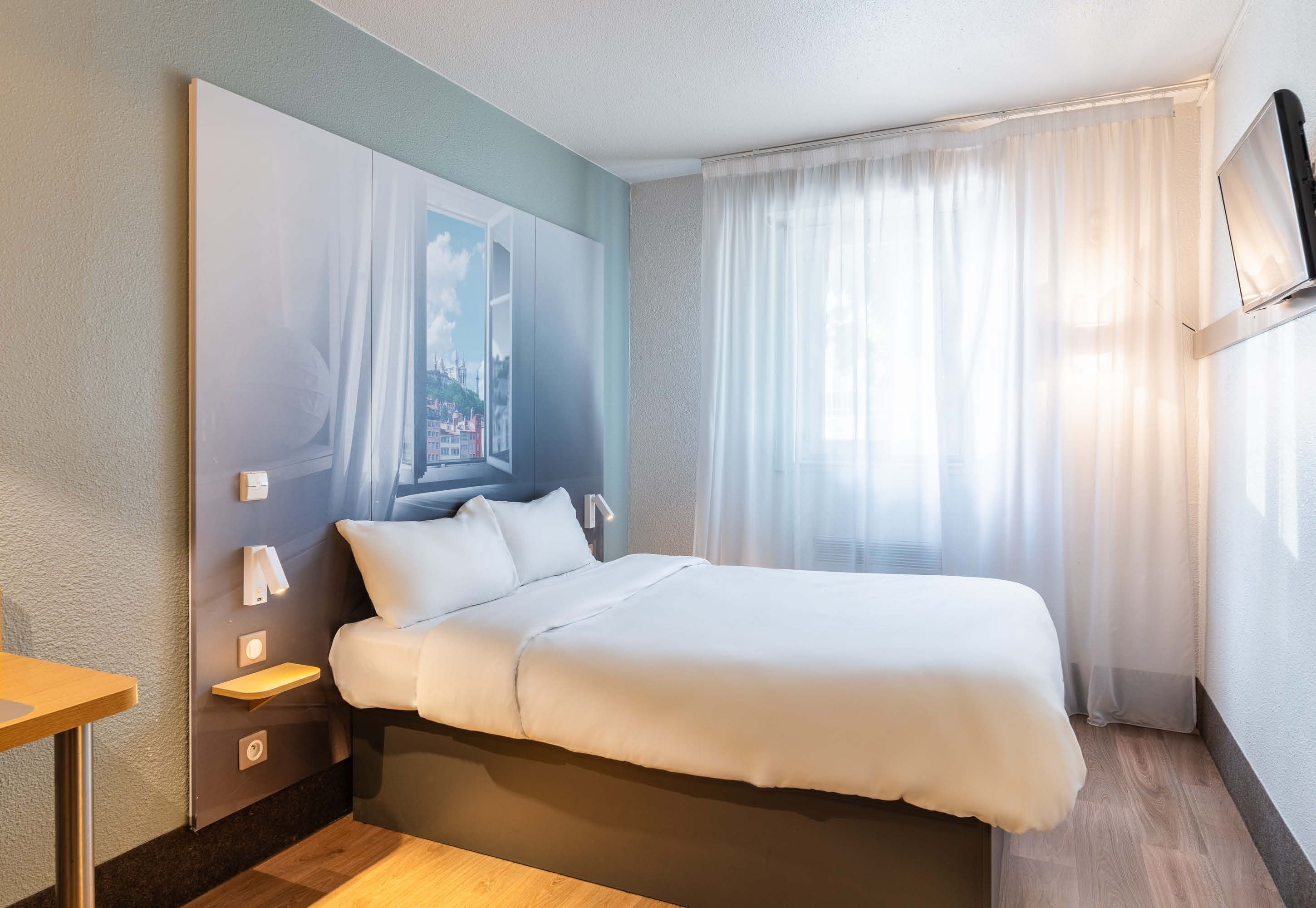 A hotel room with a bed, bedside tables, a mounted TV, and a window with curtains.
