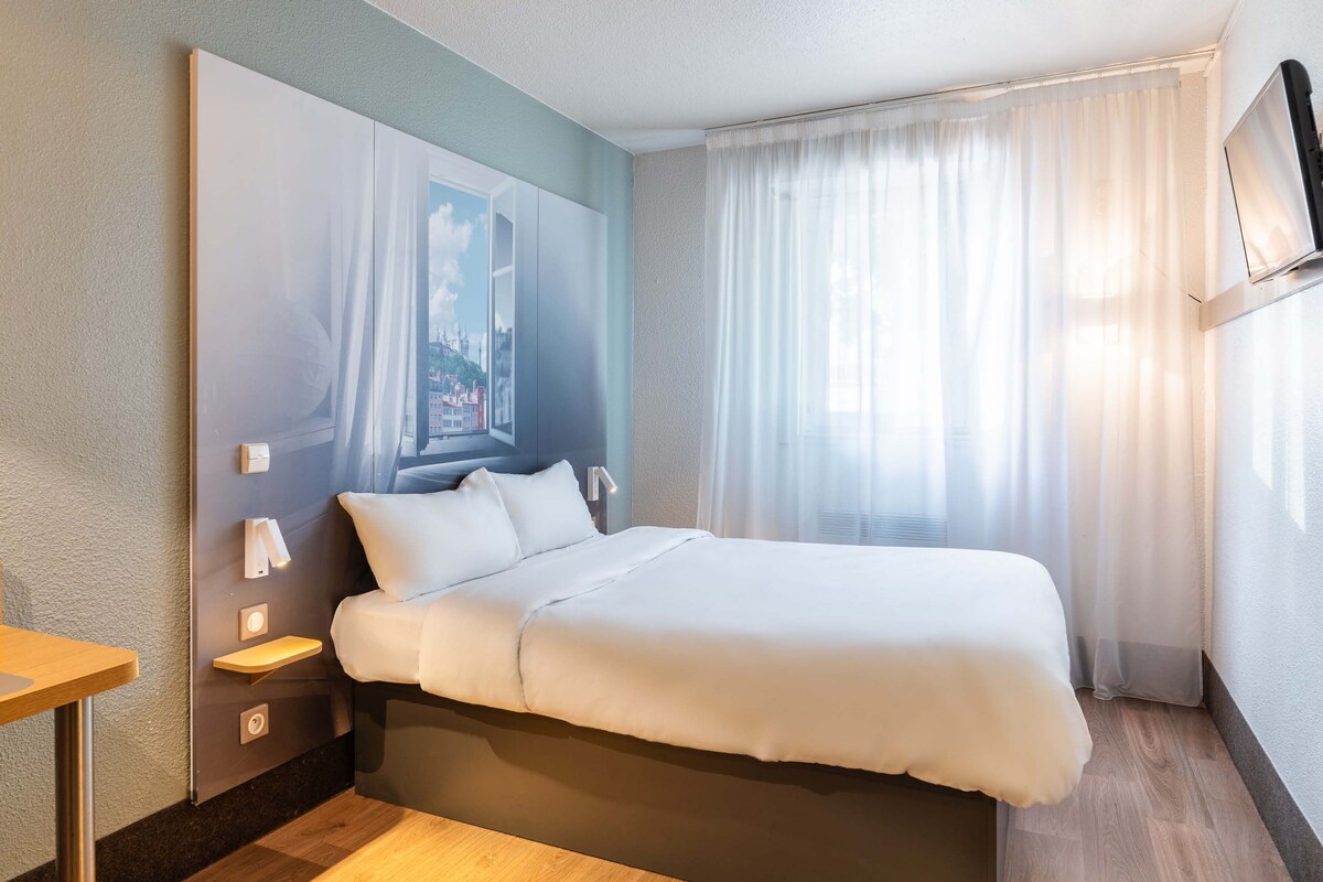 A hotel room with a bed, bedside tables, a mounted TV, and a window with curtains.