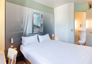 A neatly made bed with white linens, a nightstand with a lamp, and a window with a view of a cityscape. - B&B Hôtel Lyon Saint-bonnet Mi-plaine (Saint-Bonnet-de-Mure)