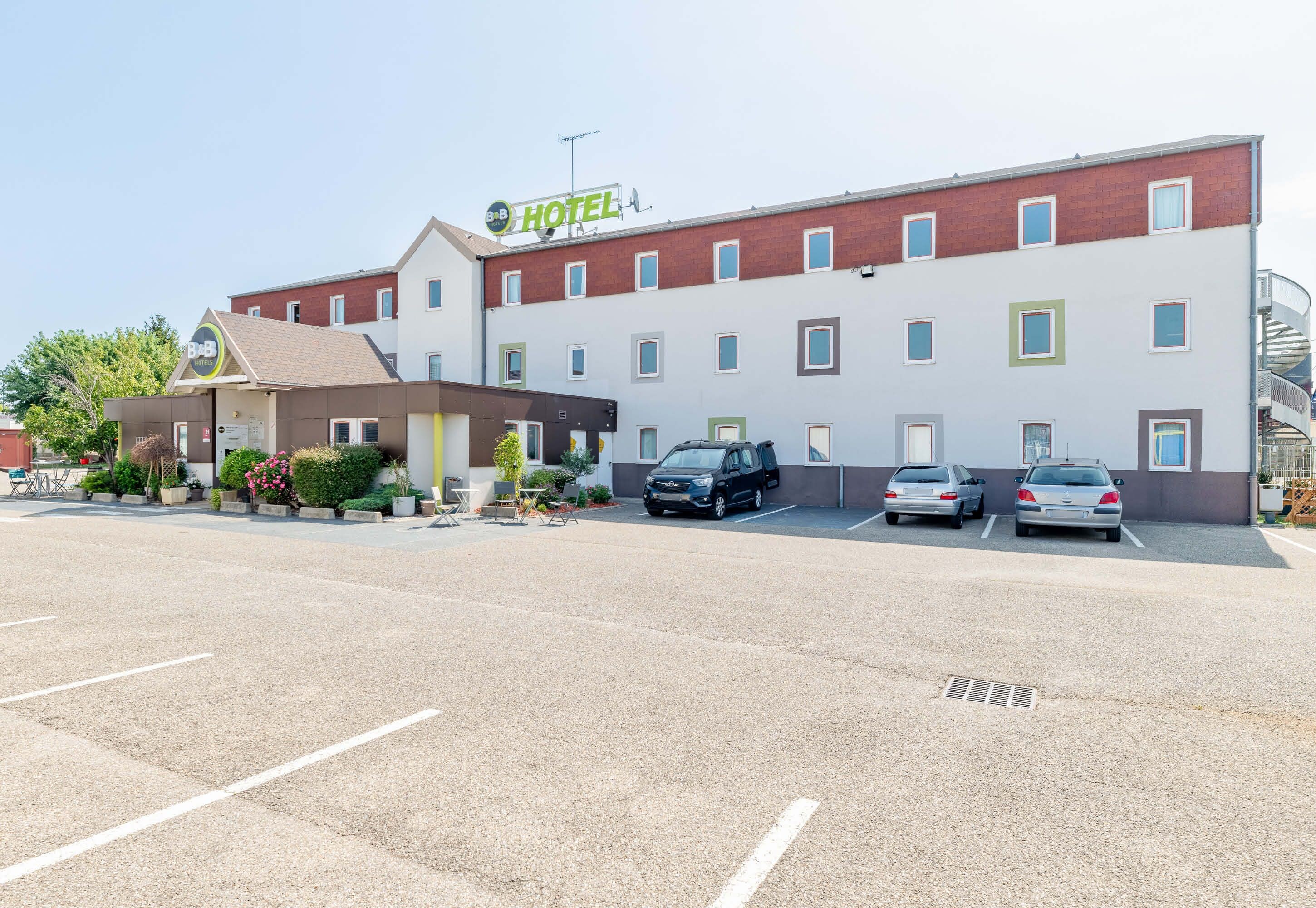 Ein Hotel im modernen Stil, mit einem Parkplatz, mehreren Autos, einer kleinen Außenbereich-Sitzgelegenheit und einem Schild mit dem Hotelnamen.